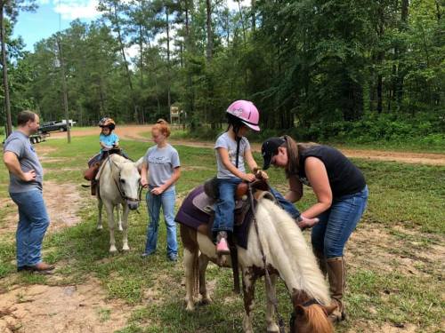 Two children on ponies, with adults assisting them in a wooded area. Everyone is smiling and enjoying the outdoor activity.