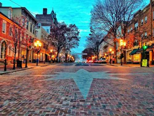 A quiet street at dusk, featuring brick pavement, trees, and charming storefronts under a blue sky.