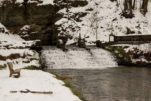 A snowy landscape featuring a small waterfall, a river, and a lone chair on the bank.