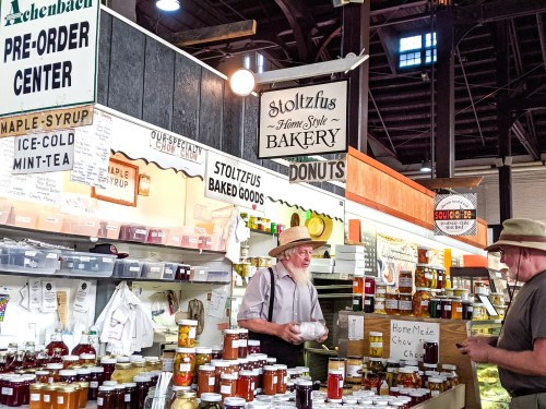 A bustling market stall featuring jars of homemade goods, with two men discussing products under colorful signs.