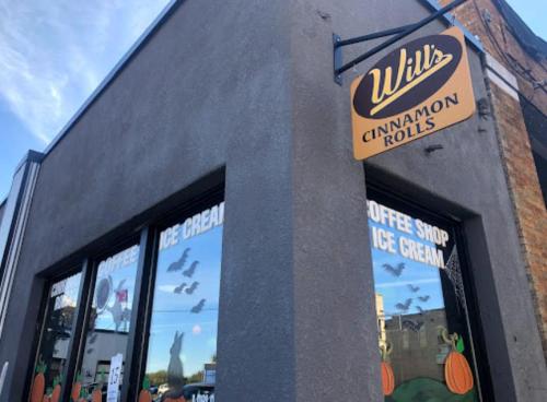 Sign for "Will's Cinnamon Rolls" on a coffee shop with windows decorated for fall, featuring pumpkins and bats.
