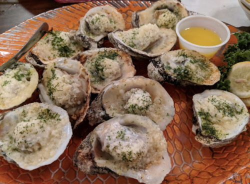 A platter of baked oysters topped with breadcrumbs and herbs, served with lemon and a small cup of dipping sauce.