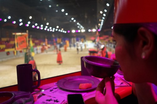 A person in a red hat watches a medieval-themed show, holding a bowl, with a colorful arena in the background.