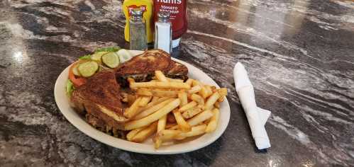 A plate with a grilled sandwich, fries, pickles, and condiments on a marble table.