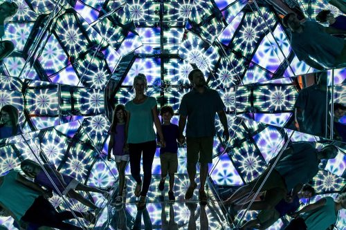A family walks through a mirrored kaleidoscope installation, surrounded by colorful patterns and reflections.