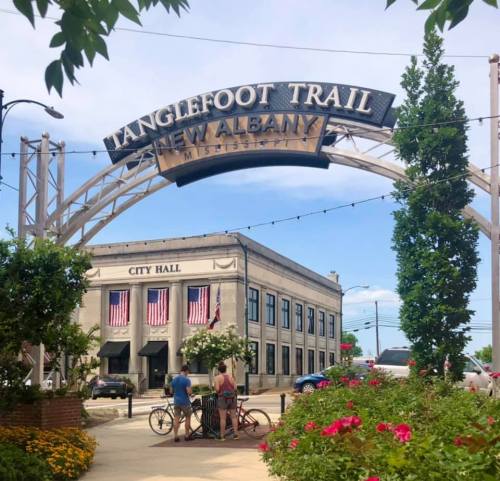 Archway sign for Tanglefoot Trail in New Albany, Mississippi, with City Hall and people enjoying the area.