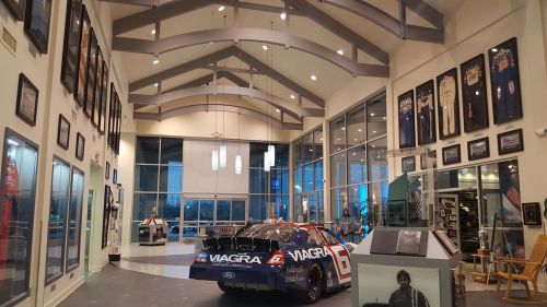A race car displayed in a spacious showroom with high ceilings, featuring memorabilia and framed jerseys on the walls.