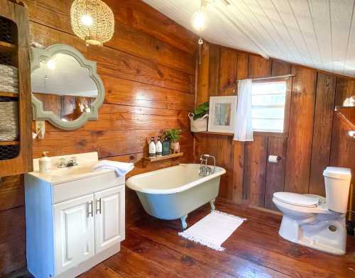 Cozy bathroom with wooden walls, a vintage tub, sink, and toilet, featuring natural light and decorative elements.