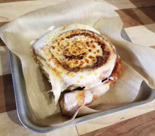 A freshly baked cinnamon roll with a golden-brown top, served in a metal tray lined with parchment paper.