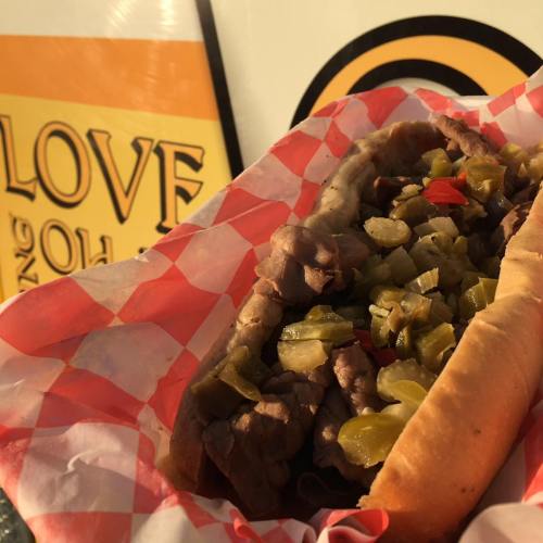 A close-up of a loaded hot dog in a checkered paper wrapper, topped with chopped onions, pickles, and peppers.