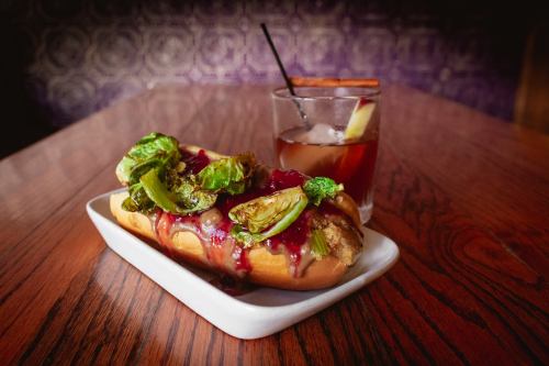 A hot dog topped with Brussels sprouts and sauce, served on a white plate next to a cocktail on a wooden table.