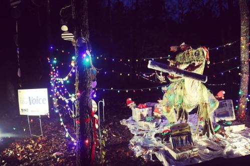 A festive dinosaur decoration surrounded by colorful lights and holiday gifts in a dark forest setting.
