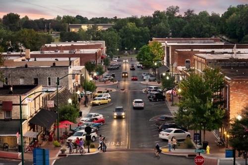 A charming small town street at dusk, lined with shops, trees, and parked cars, with people walking and dining outside.