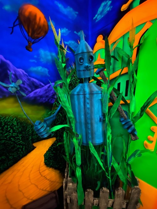 A blue tin man stands among green plants, with a colorful background featuring a yellow brick road and a hot air balloon.