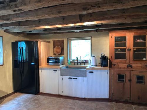 A rustic kitchen with wooden beams, a black fridge, a farmhouse sink, and a wooden cabinet. Natural light from a window.