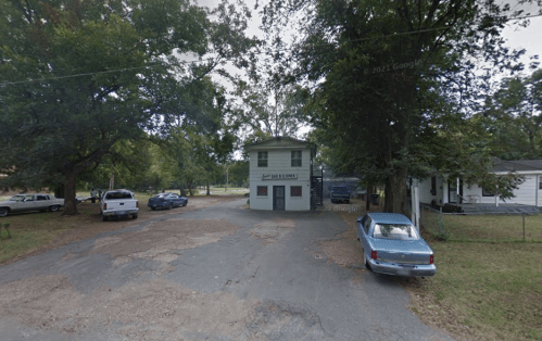 A small two-story building surrounded by trees and parked cars on a gravel lot.