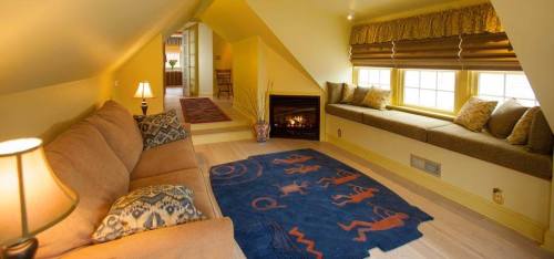 Cozy attic living space with a sofa, decorative rug, and large windows featuring a fireplace and warm yellow tones.