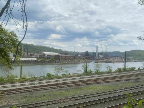 A view of a river with industrial buildings and smokestacks in the background, under a cloudy sky.