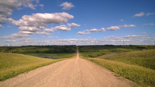 A gravel road stretches through green fields under a blue sky with fluffy clouds and wind turbines in the distance.