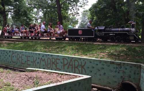 A miniature train rides through a park, with passengers enjoying the scenic view and a creek in the foreground.