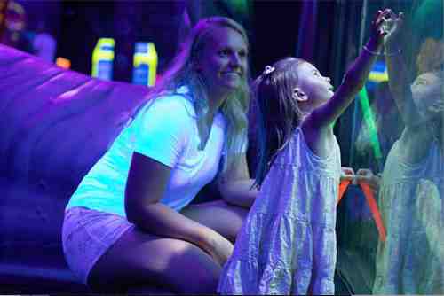 A woman smiles while a young girl reaches out to a colorful, illuminated display in a playful environment.