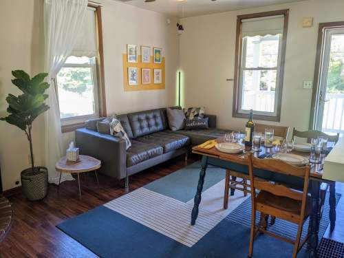 Cozy living space with a gray sofa, dining table set for dinner, and bright windows. Decor includes framed art and plants.