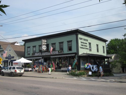 A green general store with flags, colorful merchandise outside, and people walking by on a sunny day.