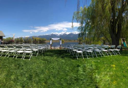 A scenic outdoor wedding setup with rows of chairs facing an altar by a lake, surrounded by mountains and greenery.