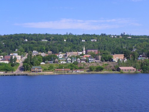 A scenic view of a riverside town with green hills, buildings, and a clear blue sky.