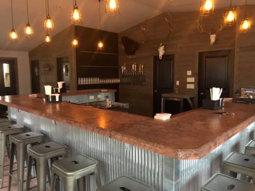 A rustic bar with a metal countertop, wooden walls, and pendant lights, featuring stools and a beer tap wall.