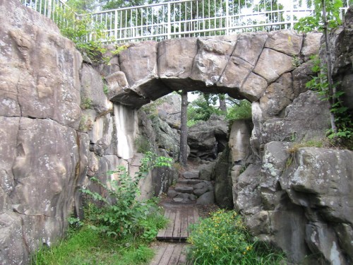 A stone archway surrounded by rocky terrain and greenery, leading to a wooded path.