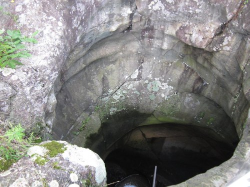 A deep, rocky well or pit surrounded by moss and vegetation, showing textured stone walls.