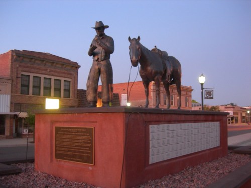 Statue of a cowboy with a horse, set on a pedestal with a plaque, in a small town at dusk.