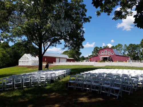 A scenic outdoor venue with white chairs set up for an event, featuring a red barn and a greenhouse in the background.