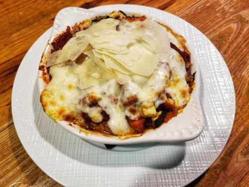 A baked dish with layers of cheese, vegetables, and sauce, topped with shaved cheese, served in a white dish on a plate.