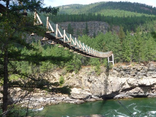 A suspension bridge stretches over a river, surrounded by lush green trees and rocky cliffs in a mountainous landscape.