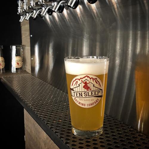 A glass of beer with a logo sits on a bar top, next to beer taps and bottles in the background.