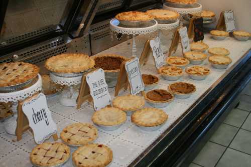 A display of various pies on stands, labeled with flavors like cherry, pecan, and pumpkin, in a bakery setting.