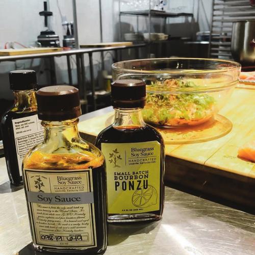 Bottles of Bluegrass Soy Sauce and Ponzu on a kitchen counter with a bowl of salad in the background.