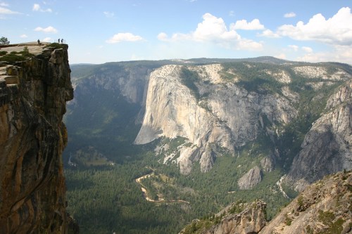 A panoramic view of a rocky cliff overlooking a lush valley and distant mountains under a partly cloudy sky.