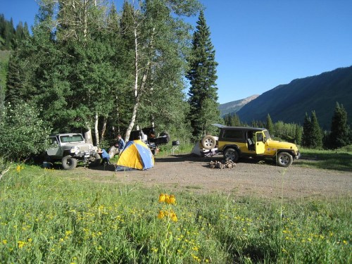 A campsite with two off-road vehicles, a yellow tent, and people enjoying the outdoors surrounded by trees and mountains.