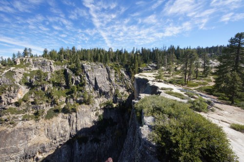 A panoramic view of a rocky cliff surrounded by lush green trees under a blue sky with scattered clouds.