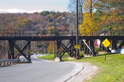 A road curves under a railway bridge, surrounded by autumn trees and warning signs.
