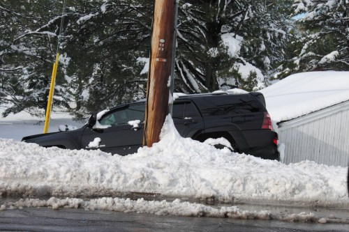 A black SUV partially buried in snow next to a utility pole and a snow-covered building.