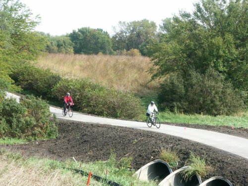 Two cyclists ride along a winding path through a grassy landscape with trees and shrubs in the background.