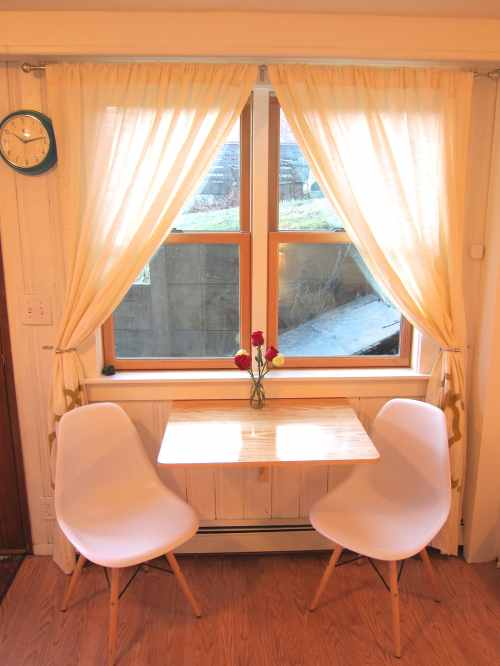 A cozy dining nook with a small table, two white chairs, and a vase of flowers by a sunny window with sheer curtains.