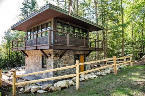 A modern two-story cabin with a stone base, large windows, and a wooden fence, surrounded by trees.
