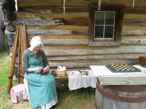 An elderly woman in period clothing sits outside a log cabin, working with yarn beside a checkers board and a table.