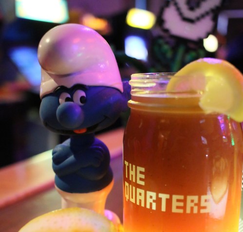 A blue cartoon character with a white hat stands next to a drink with a lemon slice, in a vibrant arcade setting.