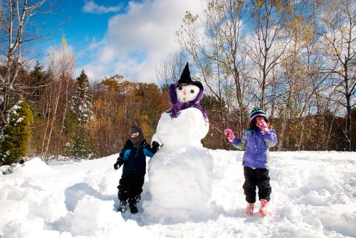 Two children play in the snow beside a snowman wearing a witch's hat and purple scarf, with trees in the background.